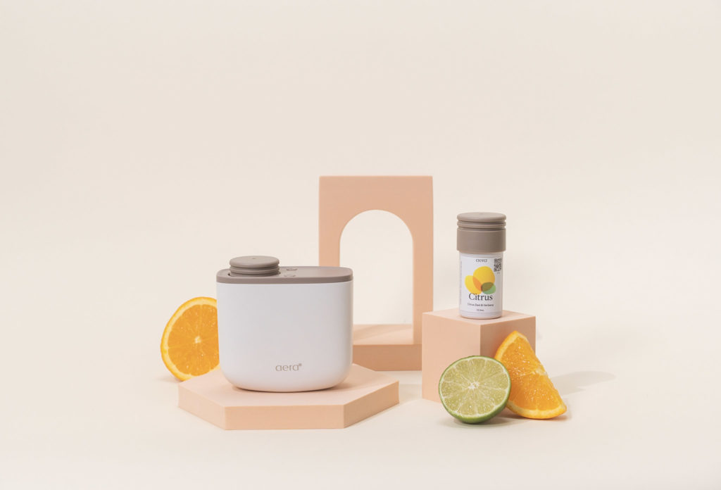 Der AERA Mini steht auf einem Podest neben dem AERA Mini Duft Citrus. Daneben liegen aufgeschnittene Orangen und eine Limette.