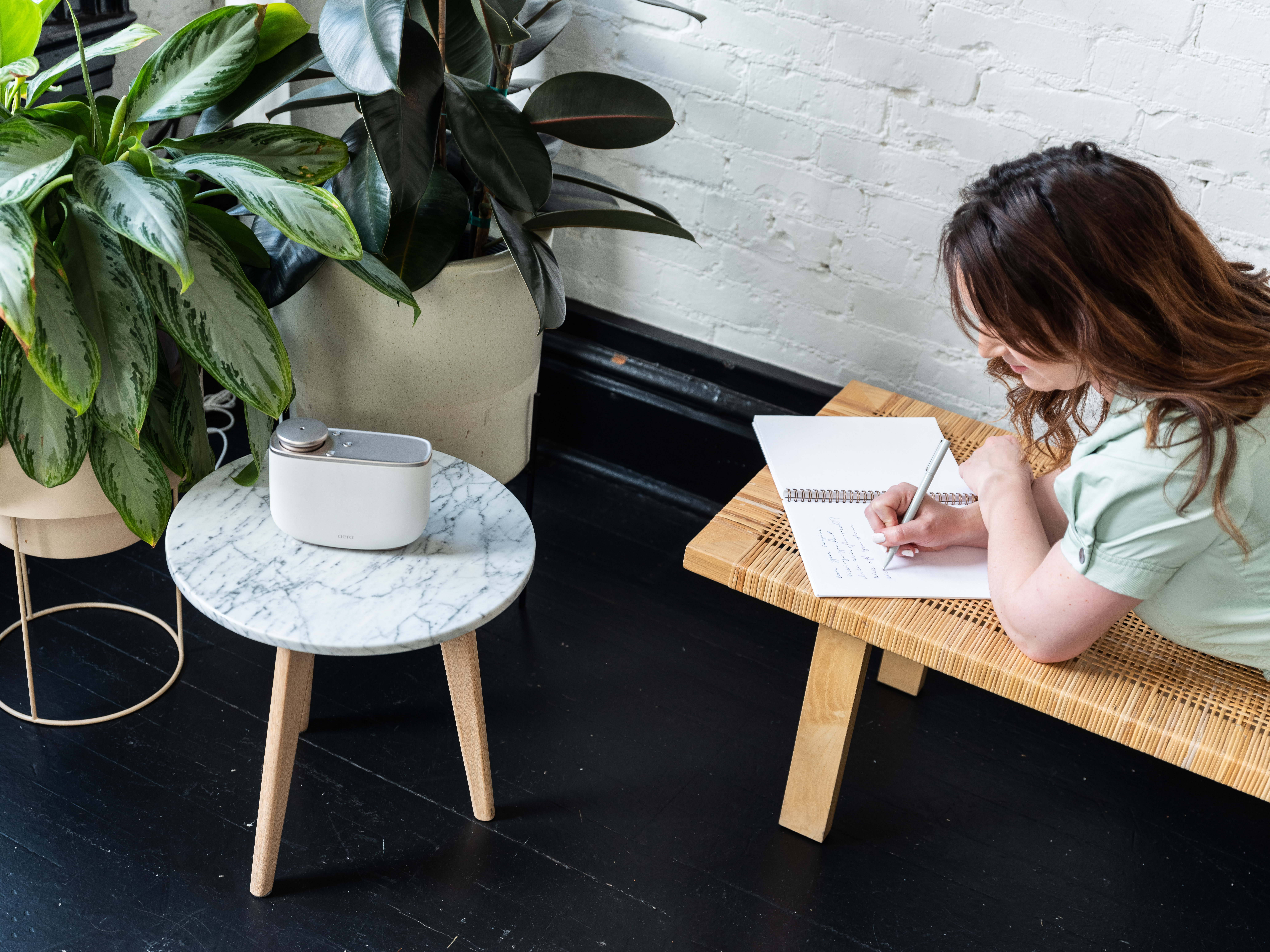 Hygge und AERA ergänzen sich. Eine Frau, die auf einer Bank liegt und schreibt. Daneben steht der AERA Smart auf einem kleinen Tisch.