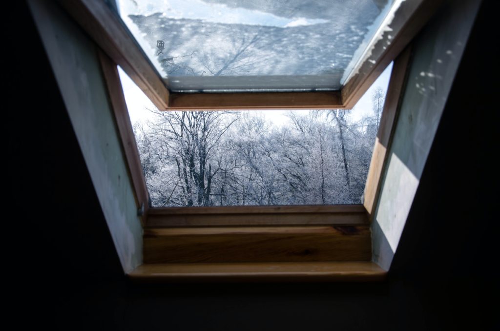 anna-dudkova-lllhioZ4tlc-unsplash Zu sehen ist en offenes Fenster in einer Dachschrägen. Der Blick geht von drinnen nach draußen auf einen schneebedeckten Wald.