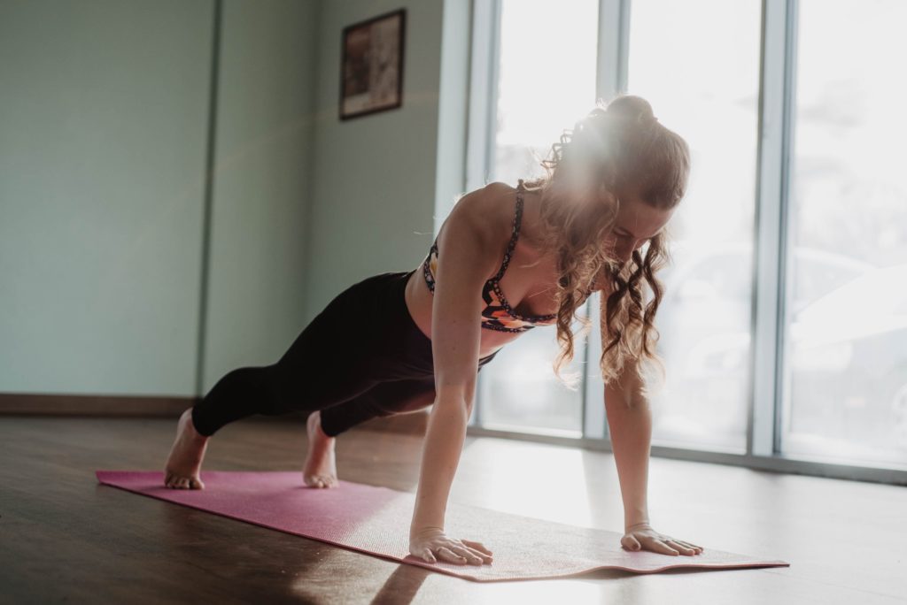 olivia-bauso-hWgsxV_VQW0-unsplash Eine junge Frau mit Locken macht den Yoga-Liegestütz auf ihrer pinken Matte, drinnen vor dem Fenster.