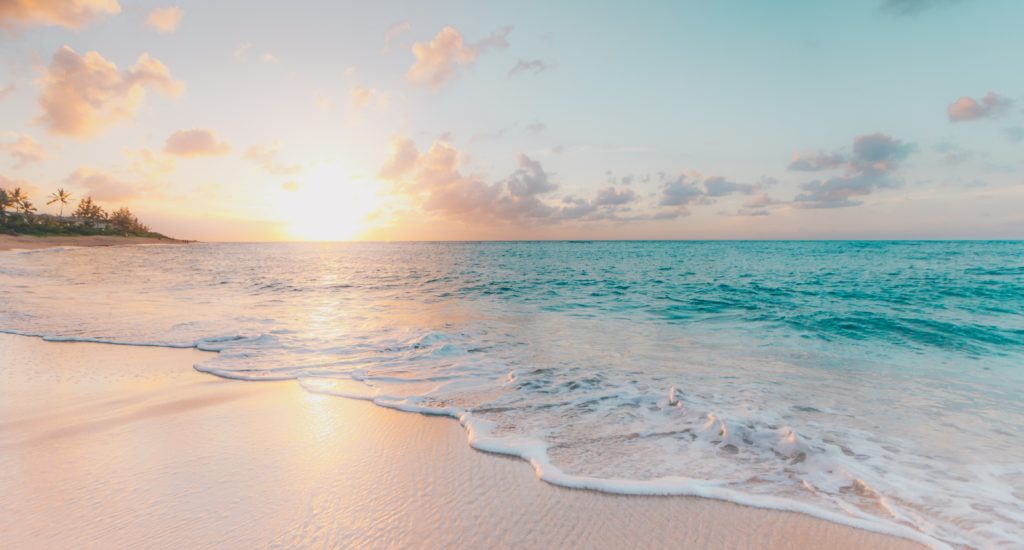 So stellen wir uns Düfte für den Sommer vor: Sandstrand mit türkisem Meer. In der Ferne sind ein paar Palmen zu sehen. Die Sonne geht zwischen ein paar Wolken unter.