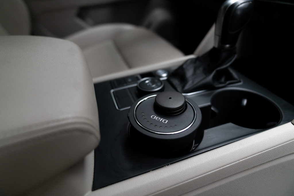 Zu sehen ist der AERA Go Car Diffuser im Getr&auml;nkehalter in der Mittelkonsole eines Autos mit grauen Ledersitzen und schwarzem Armaturenbrett.